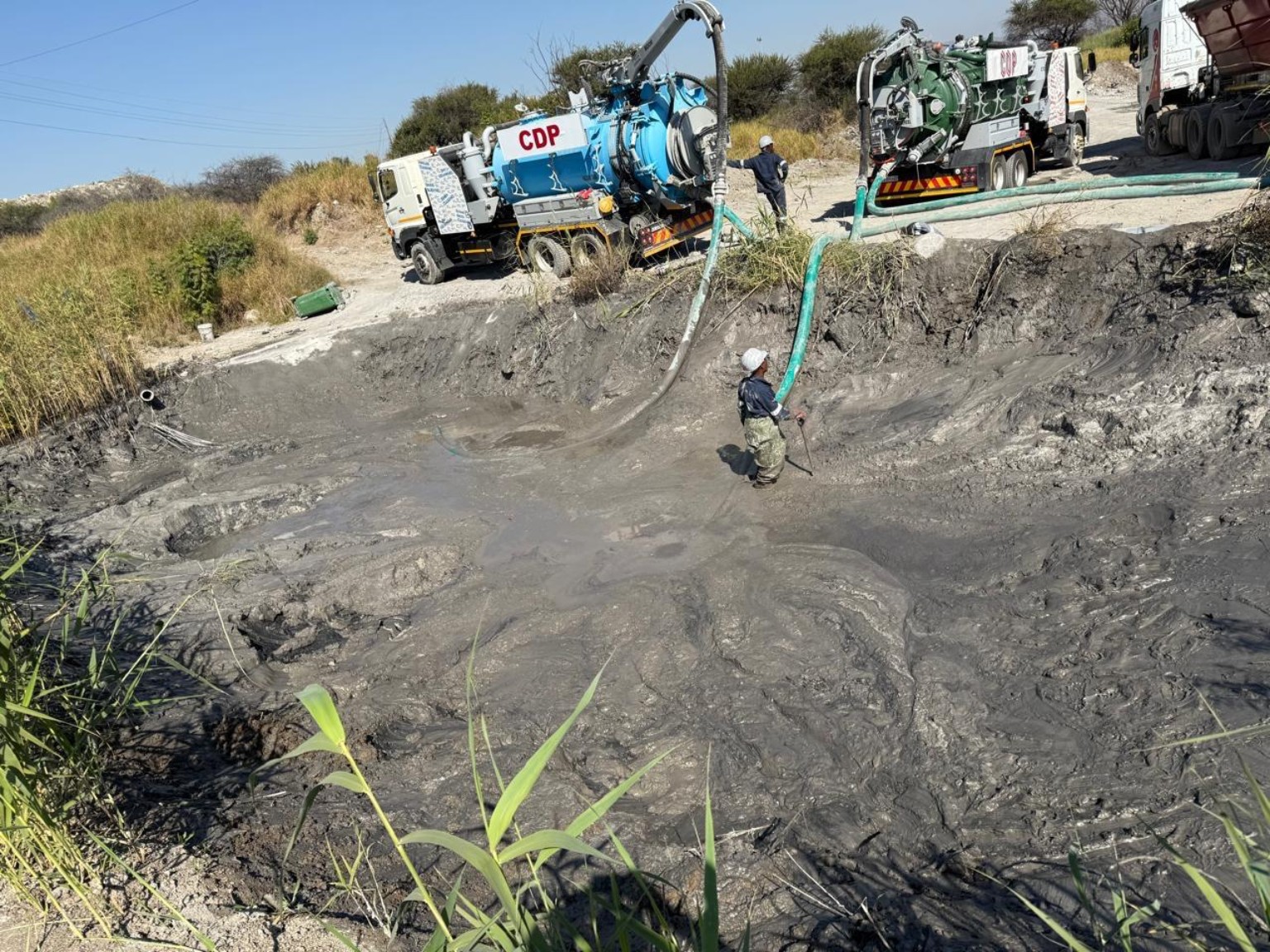 Workers handling vacuum hoses in a muddy excavation with trucks alongside